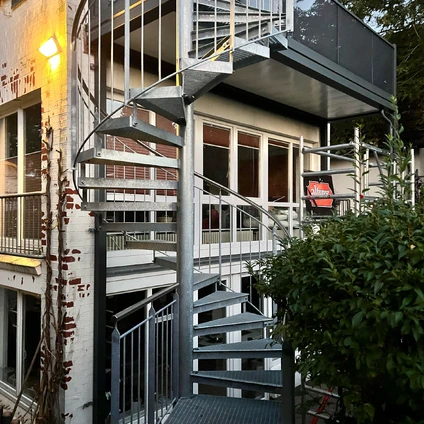Metall-Außentreppe an einer weißen Fassade flankiert von üppigem Grün und Bäumen